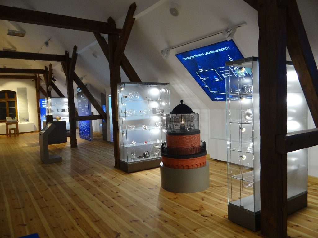 Sala ekspozycyjna Muzeum Latarnictwa. Pomiędzy przeszklonymi, podświetlanymi gablotami z eksponatami stoi miniatura szczytowej części latarni morskiej. 