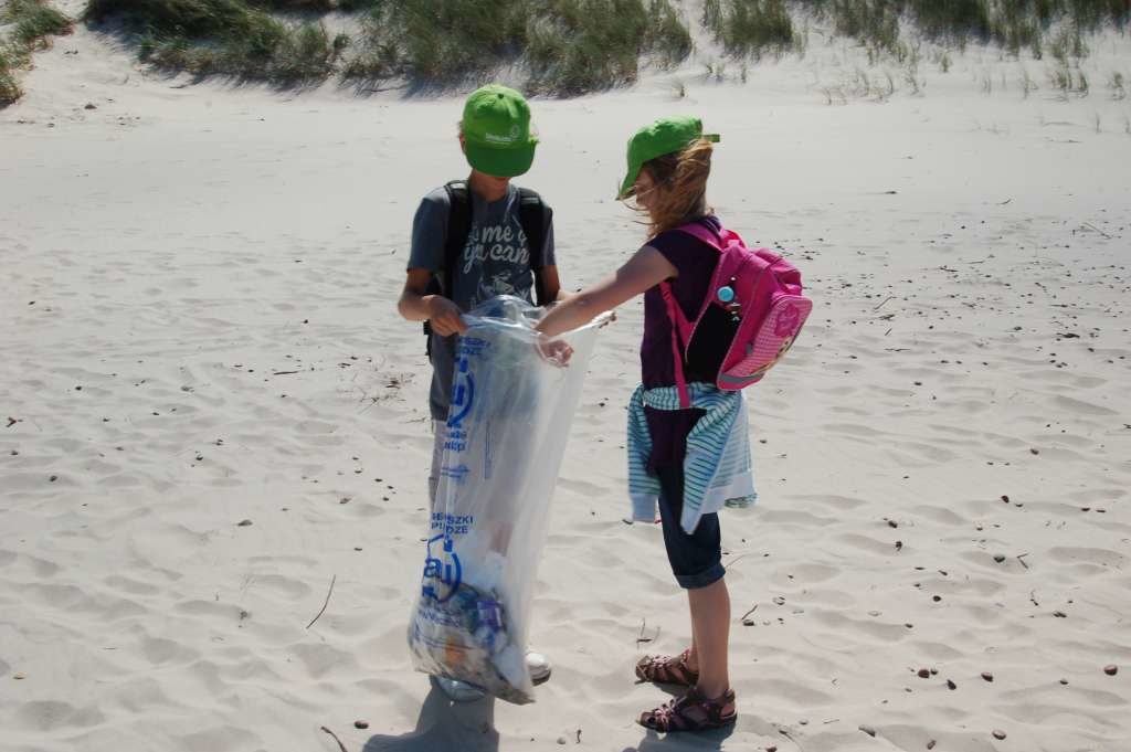 Na plaży stoi dwójka dzieci w zielonych czapeczkach. Chłopiec i dziewczynka w wieku około 10 lat trzymają między sobą duży foliowy worek z napisem alu. Worek w jednej trzeciej wypełniony jest śmieciami.