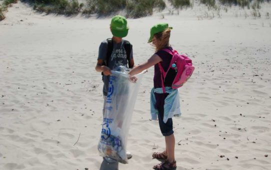 Na plaży stoi dwójka dzieci w zielonych czapeczkach. Chłopiec i dziewczynka w wieku około 10 lat trzymają między sobą duży foliowy worek z napisem alu. Worek w jednej trzeciej wypełniony jest śmieciami.