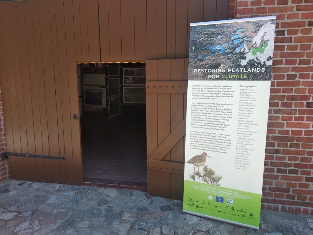 Wejście do stodoły poprzez otwarte, drewniane drzwi. Od frontu widać baner zapraszający na wystawę zdjęć, znajdującą się w środku budynku.