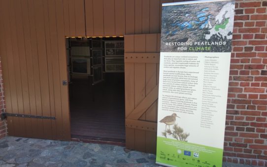 Wejście do stodoły poprzez otwarte, drewniane drzwi. Od frontu widać baner zapraszający na wystawę zdjęć, znajdującą się w środku budynku.