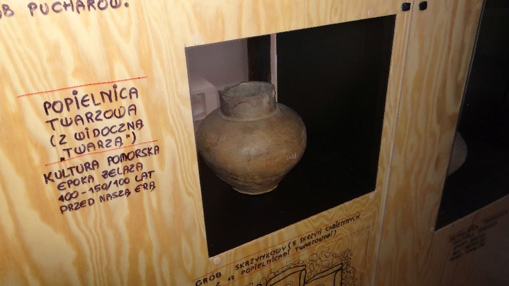 Fragment ekspozycji etnograficznej w Czołpinie. Na półce ekspozytora znajduje się gliniana, pękata popielnica pochodząca z epoki żelaza. 