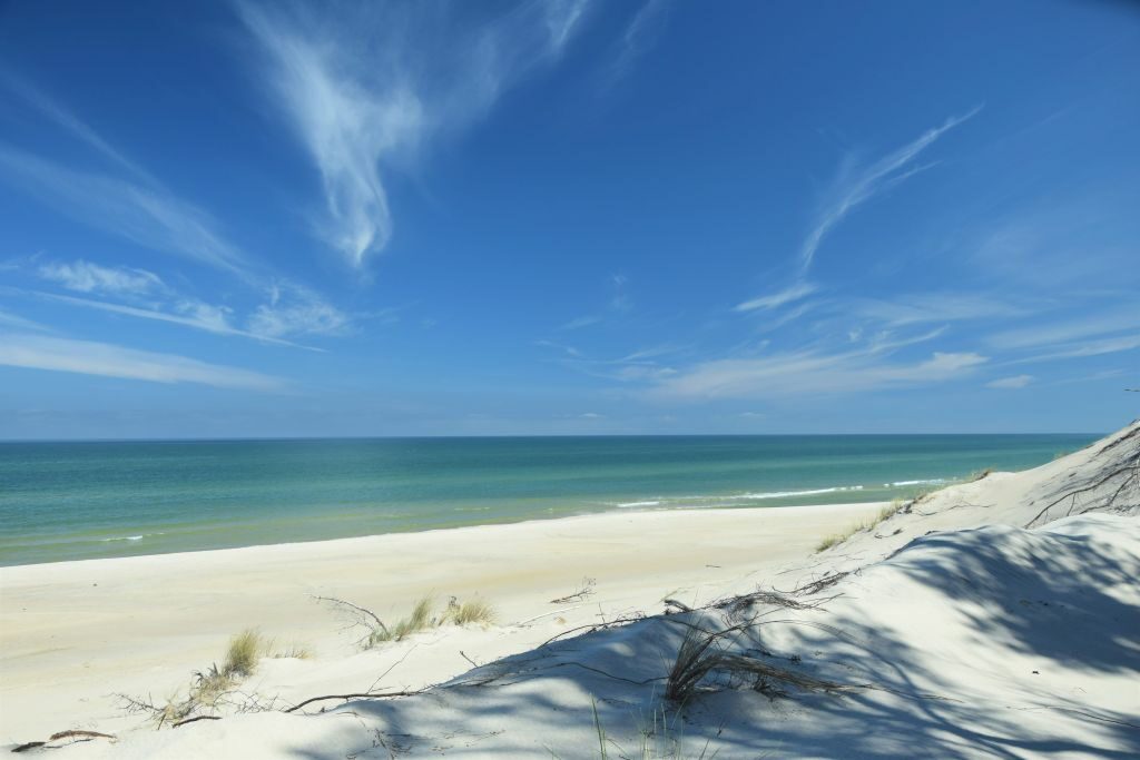 Fragment plaży i morza, biały piasek, na który pada cień z wydmy, turkusowy odcień Bałtyku, błękitne niebo, niewielkie chmury pierzaste.