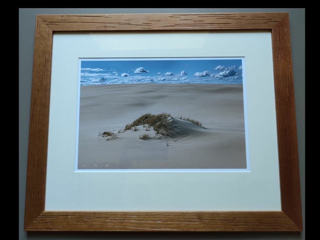 Fotografia oprawiona w drewnianą ramę przedstawiajaca małą porośnietą trawami wydmę.
