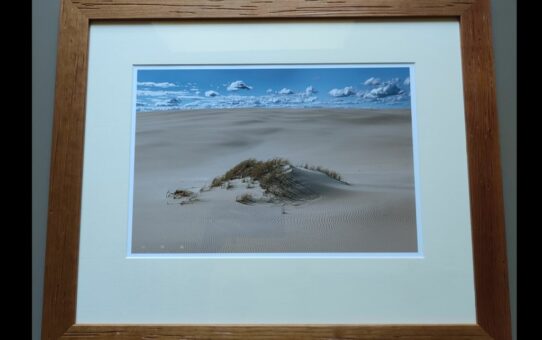 Fotografia oprawiona w drewnianą ramę przedstawiajaca małą porośnietą trawami wydmę.